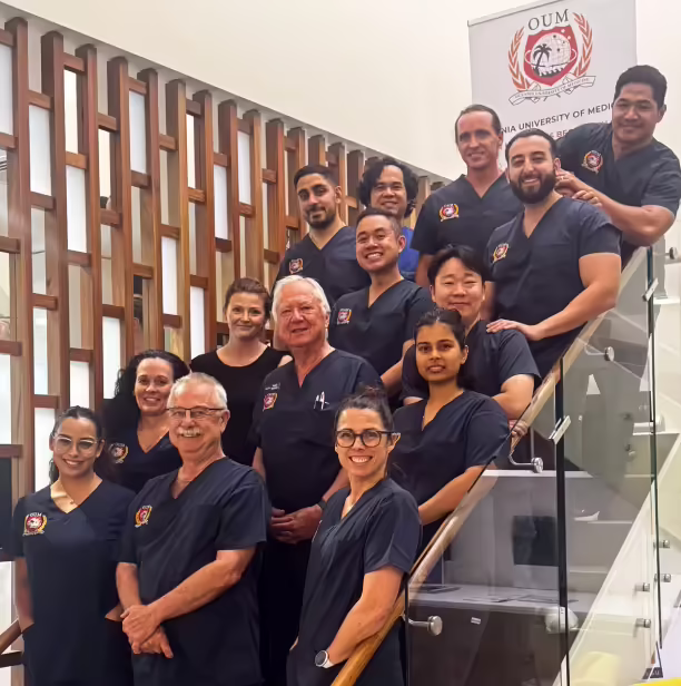 A group of doctors, wearing dark blue scrubs, pose on a glass staircase. The setting appears to be a hospital or medical facility.