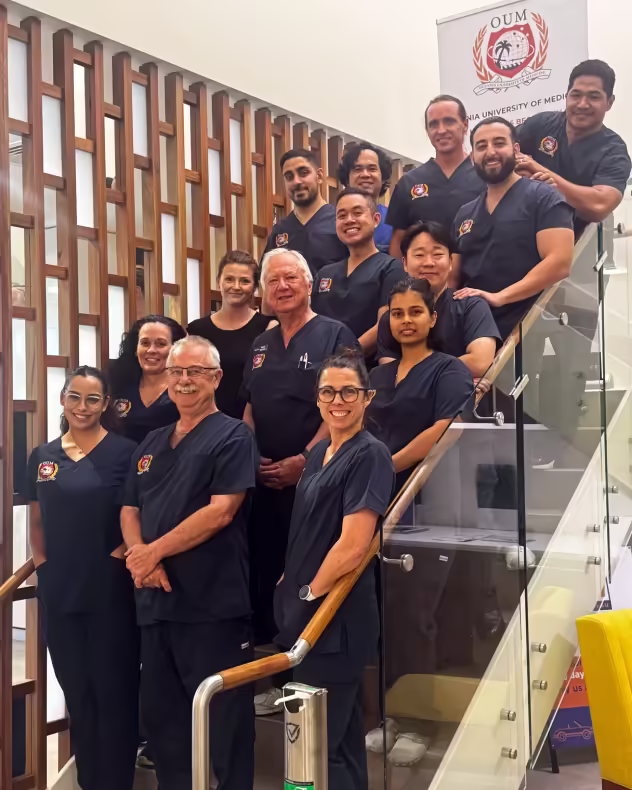 A group of doctors, wearing dark blue scrubs, pose on a glass staircase. The setting appears to be a hospital or medical facility.